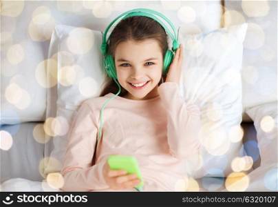people, children and technology concept - happy smiling girl lying awake with smartphone and headphones in bed listening to music over holidays lights background. girl with headphones listening to music in bed