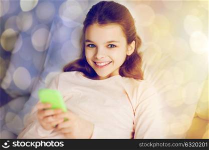 people, children and technology concept - happy smiling girl lying awake with smartphone in bed over holidays lights background. happy girl in bed with smartphone over lights