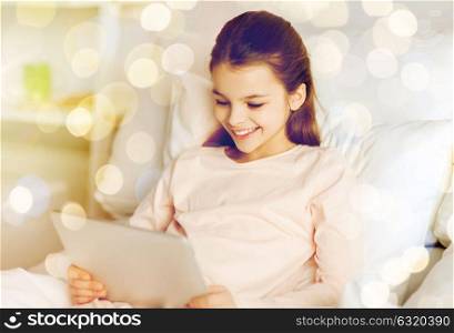 people, children and technology concept - happy smiling girl lying awake with tablet pc computer in bed at home over holidays lights background. happy girl in bed with tablet pc over lights