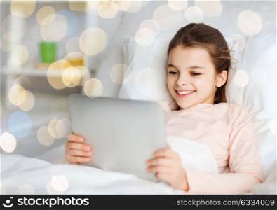 people, children and technology concept - happy smiling girl lying awake with tablet pc computer in bed at home over holidays lights background. happy girl in bed with tablet pc over lights