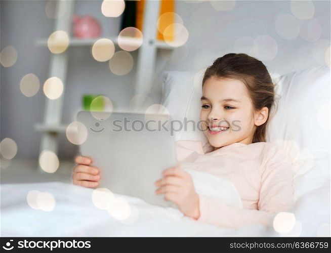 people, children and technology concept - happy smiling girl lying awake with tablet pc computer in bed at home over holidays lights. happy girl lying in bed with tablet pc at home