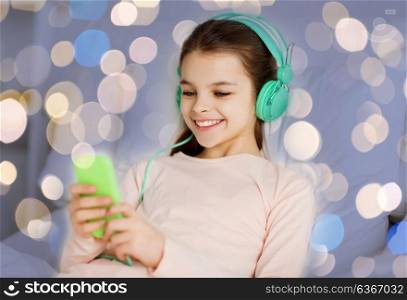 people, children and technology concept - happy smiling girl lying awake with smartphone and headphones in bed listening to music at home over holidays lights. girl in headphones with smartphone over lights