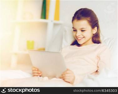 people, children and technology concept - happy smiling girl with tablet pc computer lying in bed at home. happy girl with tablet pc lying in bed at home