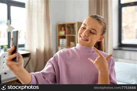 people, children and technology concept - happy smiling teenage girl with smartphone taking selfie and showing rock hand sign at home. happy girl with smartphone taking selfie at home