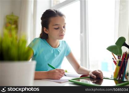 people, children, education and learning concept - girl with tablet pc computer doing homework and writing to notebook at home. girl with tablet pc writing to notebook at home. girl with tablet pc writing to notebook at home