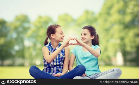 people, children, friends and friendship concept - happy little girls sitting and showing heart shape hand sign over summer park background
