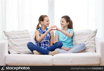 people, children, friends and friendship concept - happy little girls sitting on sofa and showing heart shape hand sign at home