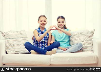 people, children, friends and friendship concept - happy little girls sitting on sofa and showing heart shape hand sign at home