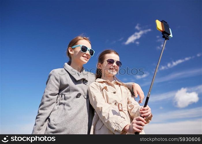 people, children, friends and friendsip concept - happy girls taking picture with smartphone on selfie stick outdoors