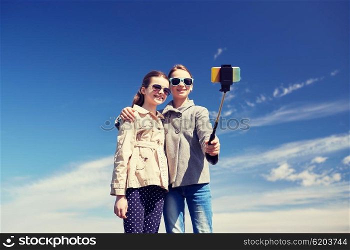 people, children, friends and friendsip concept - happy girls taking picture with smartphone on selfie stick outdoors