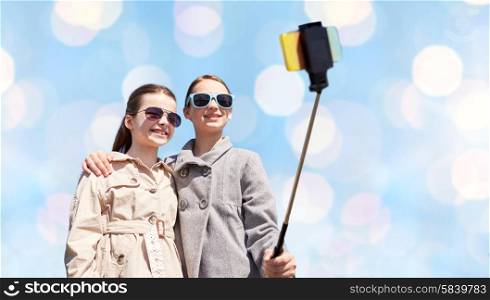people, children, friends and technology concept - happy girls taking picture with smartphone on selfie stick over blue holidays lights background