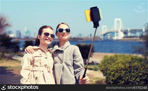 people, children, friends and technology concept - happy girls taking picture with smartphone on selfie stick over tokyo rainbow bridge in japan background