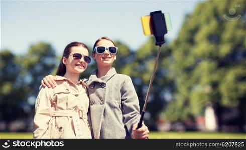 people, children, friends and technology concept - happy girls taking picture with smartphone on selfie stick over park background
