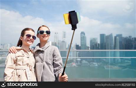 people, children, friends and technology concept - happy girls taking picture with smartphone on selfie stick over in singapore city background