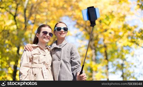 people, children, friendship and season concept - happy girls taking picture with smartphone on selfie stick over autumn park background