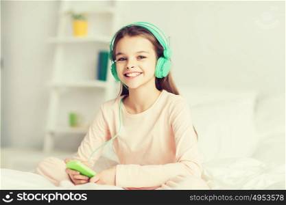 people, children, pajama party and technology concept - happy smiling girl in headphones sitting on bed with smartphone and listening to music at home. girl sitting on bed with smartphone and headphones
