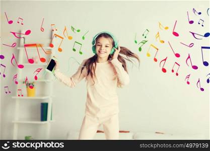 people, children, pajama party and technology concept - happy smiling girl in headphones jumping on bed with smartphone and listening to music at home. girl jumping on bed with smartphone and headphones