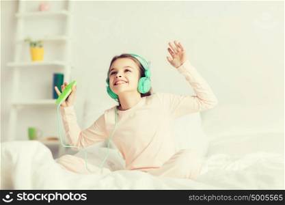 people, children, pajama party and technology concept - happy smiling girl in headphones sitting on bed with smartphone and listening to music at home. girl sitting on bed with smartphone and headphones
