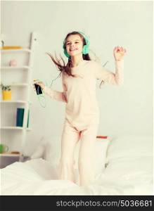 people, children, pajama party and technology concept - happy smiling girl in headphones jumping on bed with smartphone and listening to music at home. girl jumping on bed with smartphone and headphones