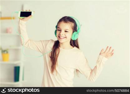 people, children, pajama party and technology concept - happy smiling girl in headphones with smartphone and listening to music at home. girl with smartphone and headphones at home