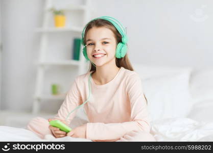 people, children, pajama party and technology concept - happy smiling girl in headphones sitting on bed with smartphone and listening to music at home