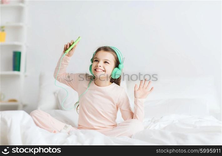 people, children, pajama party and technology concept - happy smiling girl in headphones sitting on bed with smartphone and listening to music at home