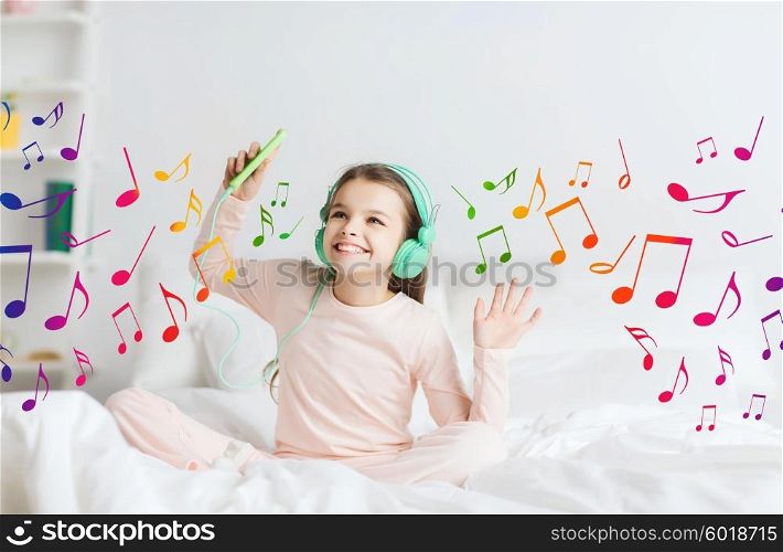 people, children, pajama party and technology concept - happy smiling girl in headphones sitting on bed with smartphone and listening to music at home over musical notes
