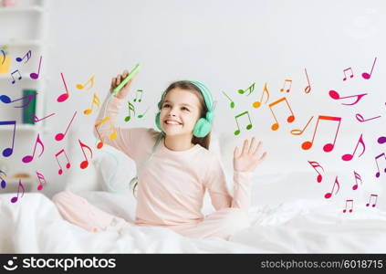 people, children, pajama party and technology concept - happy smiling girl in headphones sitting on bed with smartphone and listening to music at home over musical notes