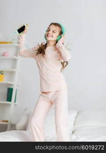 people, children, pajama party and technology concept - happy smiling girl in headphones jumping on bed with smartphone and listening to music at home