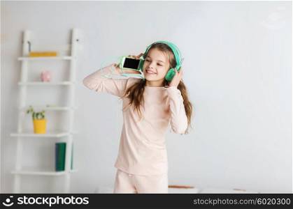 people, children, pajama party and technology concept - happy smiling girl in headphones jumping on bed with smartphone and listening to music at home