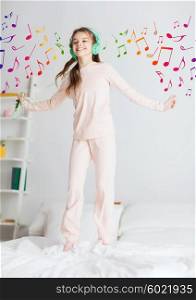 people, children, pajama party and technology concept - happy smiling girl in headphones jumping on bed with smartphone and listening to music at home over musical notes