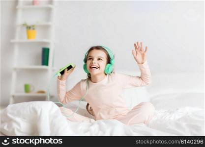 people, children, pajama party and technology concept - happy smiling girl in headphones sitting on bed with smartphone and listening to music at home
