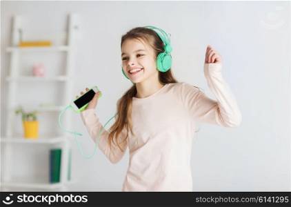 people, children, pajama party and technology concept - happy smiling girl in headphones with smartphone and listening to music at home