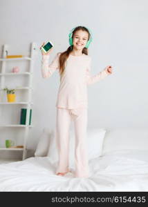 people, children, pajama party and technology concept - happy smiling girl in headphones jumping on bed with smartphone and listening to music at home