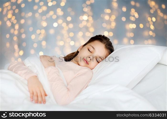 people, children, rest and comfort concept - girl sleeping in bed over holidays lights background. girl sleeping in bed over holidays lights