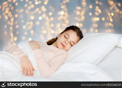 people, children, rest and comfort concept - girl sleeping in bed over holidays lights background. girl sleeping in bed over holidays lights