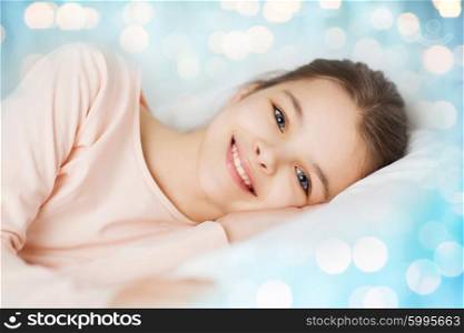 people, children, rest and comfort concept - happy smiling girl lying awake in bed over blue lights background