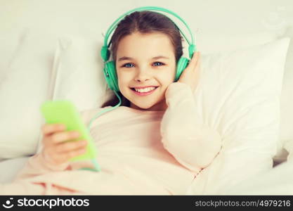 people, children, rest, and technology concept - happy smiling girl lying awake with smartphone and headphones in bed listening to music at home. happy girl lying in bed with smartphone at home