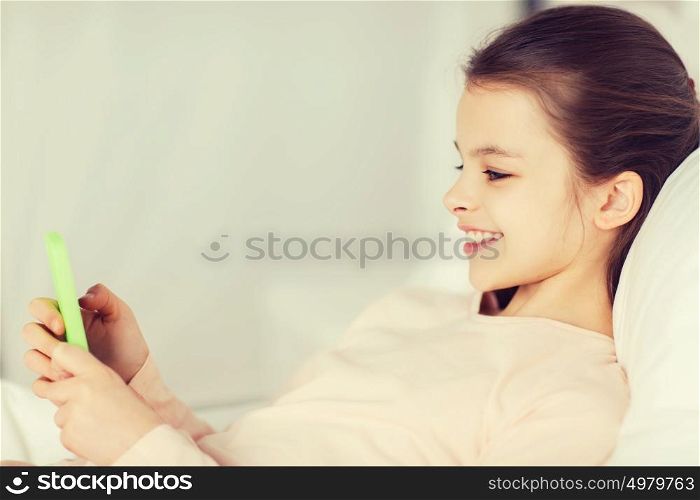 people, children, rest and technology concept - happy smiling girl lying awake with smartphone in bed at home. happy girl lying in bed with smartphone at home