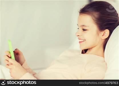 people, children, rest and technology concept - happy smiling girl lying awake with smartphone in bed at home. happy girl lying in bed with smartphone at home
