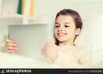 people, children, rest and technology concept - happy smiling girl lying awake with tablet pc computer in bed at home. happy girl lying in bed with tablet pc at home