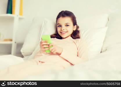 people, children, rest and technology concept - happy smiling girl lying awake with smartphone in bed at home. happy girl lying in bed with smartphone at home