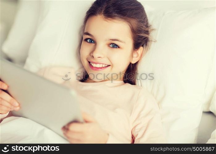 people, children, rest and technology concept - happy smiling girl lying awake with tablet pc computer in bed at home. happy girl lying in bed with tablet pc at home
