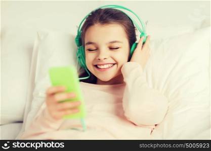 people, children, rest, and technology concept - happy smiling girl lying awake with smartphone and headphones in bed listening to music at home. happy girl lying in bed with smartphone at home