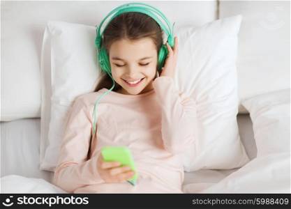 people, children, rest, and technology concept - happy smiling girl lying awake with smartphone and headphones in bed listening to music at home