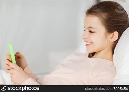 people, children, rest and technology concept - happy smiling girl lying awake with smartphone in bed at home