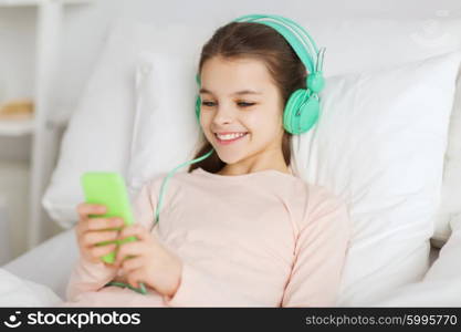 people, children, rest, and technology concept - happy smiling girl lying awake with smartphone and headphones in bed listening to music at home