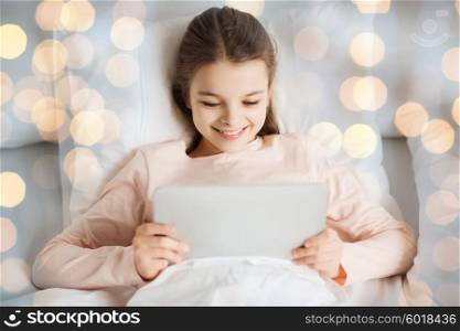 people, children, rest and technology concept - happy smiling girl lying awake with tablet pc computer in bed over lights