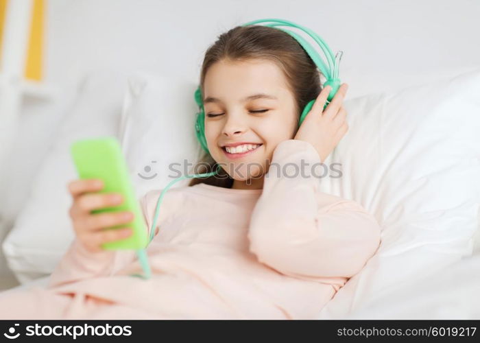 people, children, rest, and technology concept - happy smiling girl lying awake with smartphone and headphones in bed listening to music at home