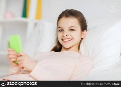 people, children, rest and technology concept - happy smiling girl lying awake with smartphone in bed at home